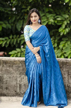 Woman wearing a blue saree with a green and white blouse against a leafy background
