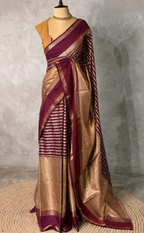 A purple and gold silk saree displayed on a mannequin against a textured gray wall. The saree features a deep purple border, a gold and purple striped body, and a solid gold pallu with small gold motifs.