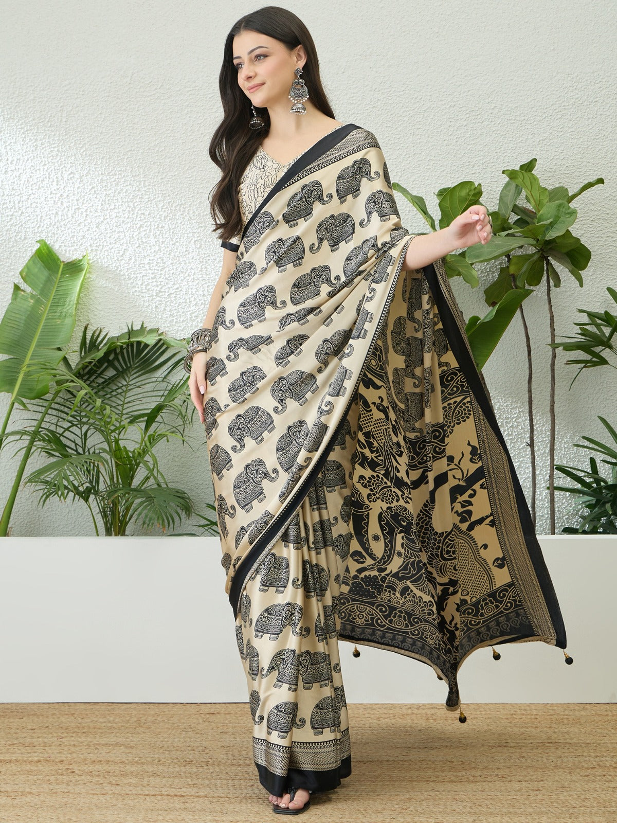 Woman wearing a black and beige saree with elephant patterns against a white wall with plants.
