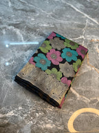 A folded patterned fabric or scarf with a black base, colorful floral motifs in pink, teal, and green, and a metallic gold border, displayed on a gray marble surface.