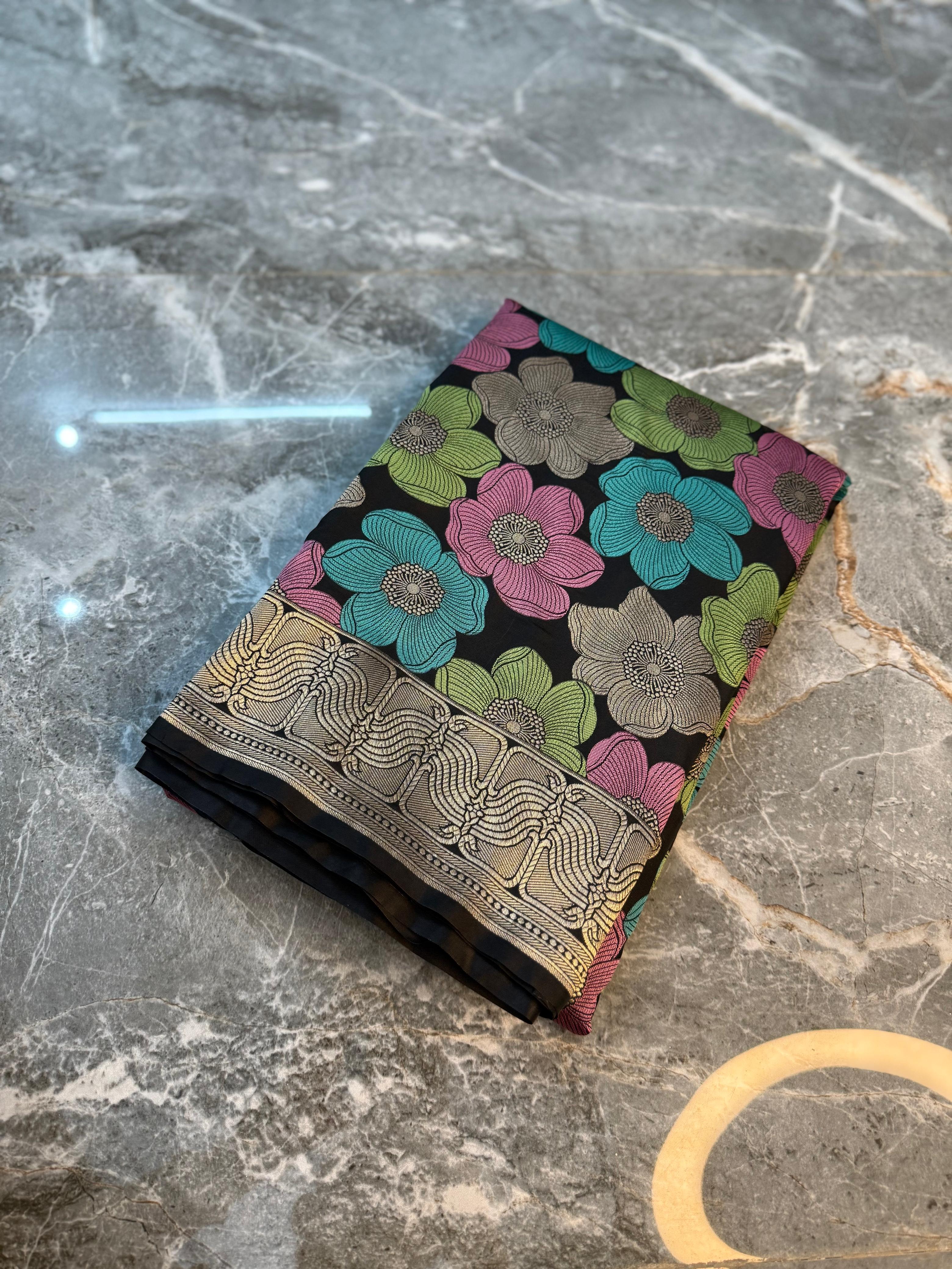A folded patterned fabric or scarf with a black base, colorful floral motifs in pink, teal, and green, and a metallic gold border, displayed on a gray marble surface.