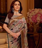 A woman wearing a traditional Indian saree in a warm indoor setting. The saree has a muted sage green base with a floral pattern in dusty pink, lavender, and black, accented by a wide metallic gold border.