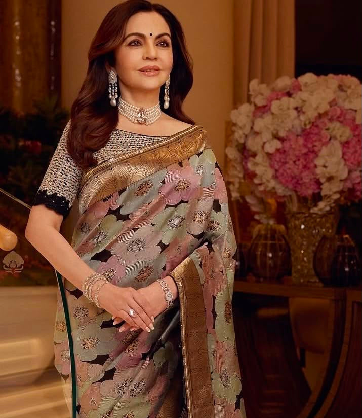 A woman wearing a traditional Indian saree in a warm indoor setting. The saree has a muted sage green base with a floral pattern in dusty pink, lavender, and black, accented by a wide metallic gold border.