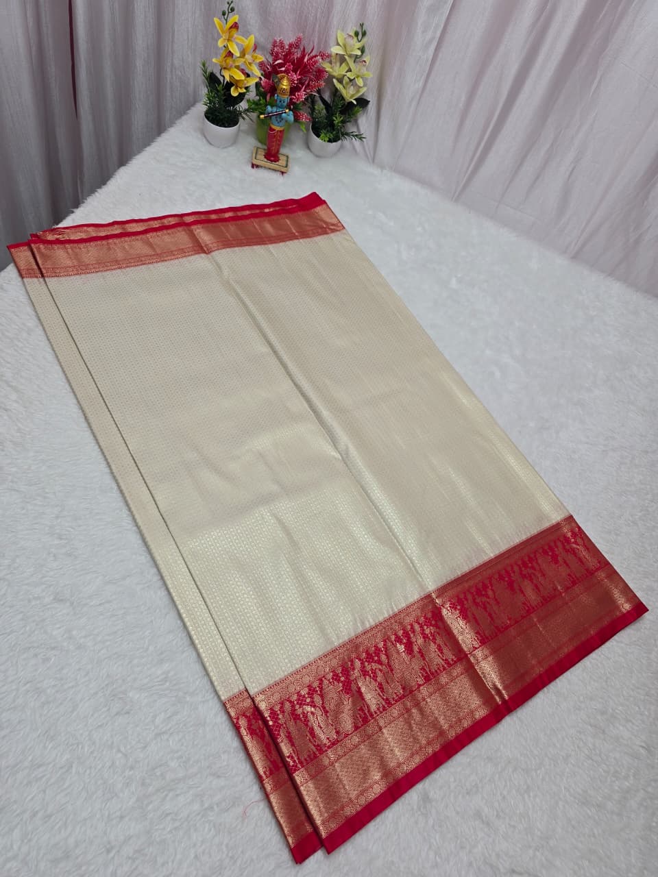 Beige saree with red border on a white surface, flowers in the background