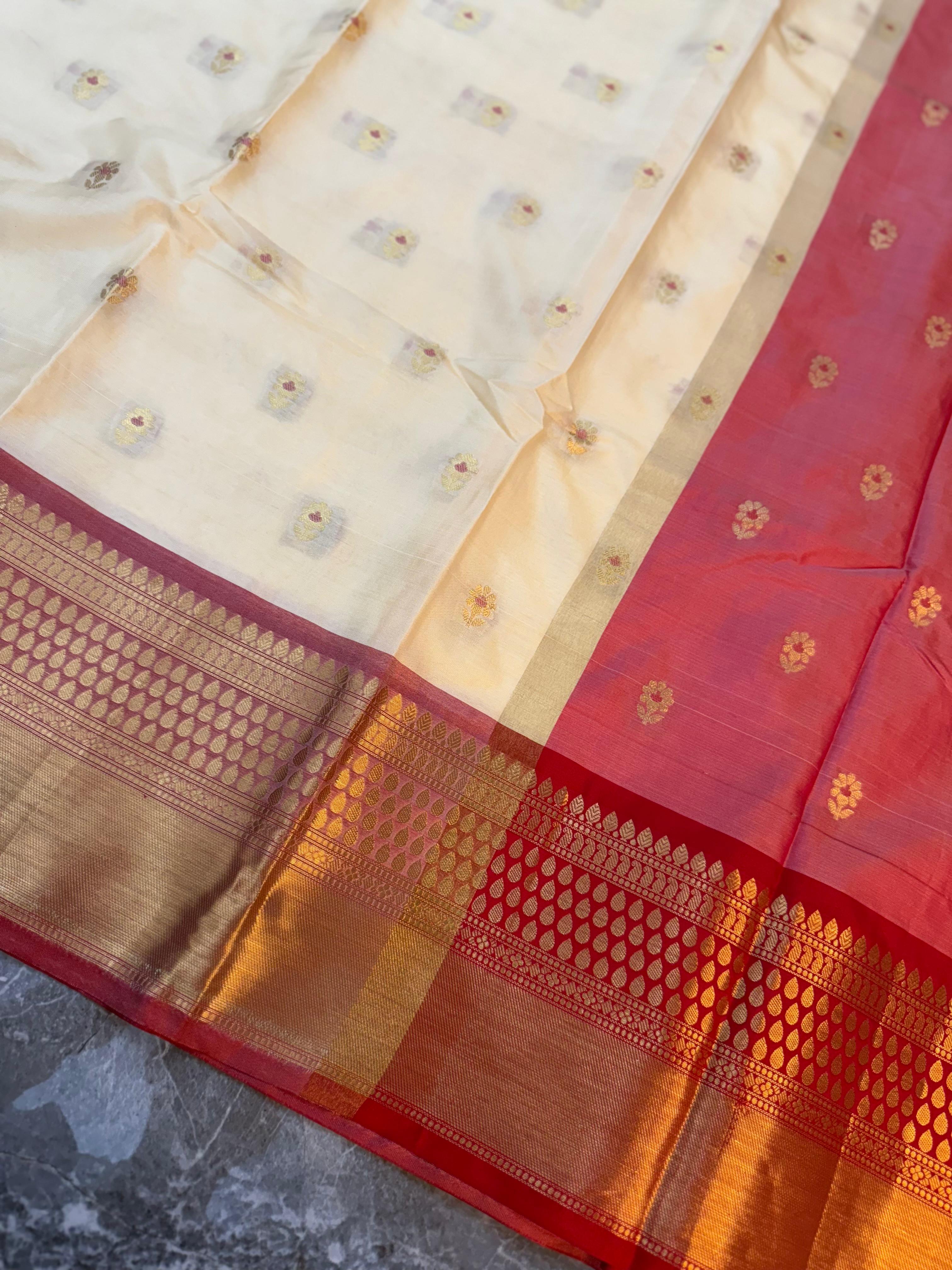 A close-up of a traditional Indian saree, showing a cream-colored body with small floral motifs and a rich red border accented by a wide gold zari brocade.