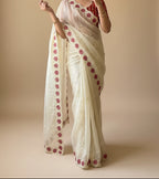 Woman wearing a white saree with red patterns in a room with plants.