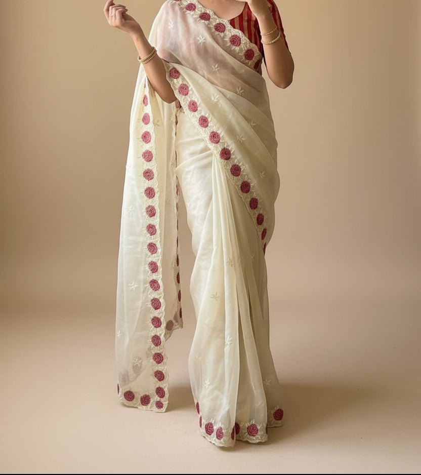 Woman wearing a white saree with red patterns in a room with plants.
