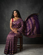 Woman in a purple saree with gold patterns sitting on a chair against a gray background