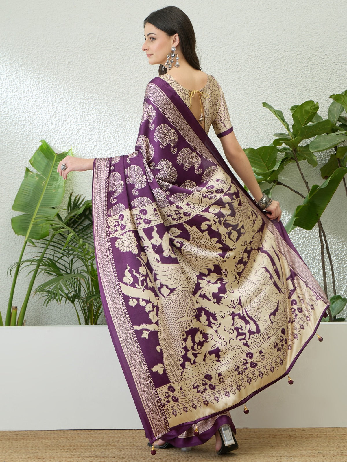 Woman wearing a purple and beige saree with elephant patterns, standing indoors with plants in the background.