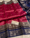 A folded and draped magenta pink silk saree with metallic gold zari patterns, a navy blue border, and a matching navy blue and gold pallu, with a small price card placed on the fabric.