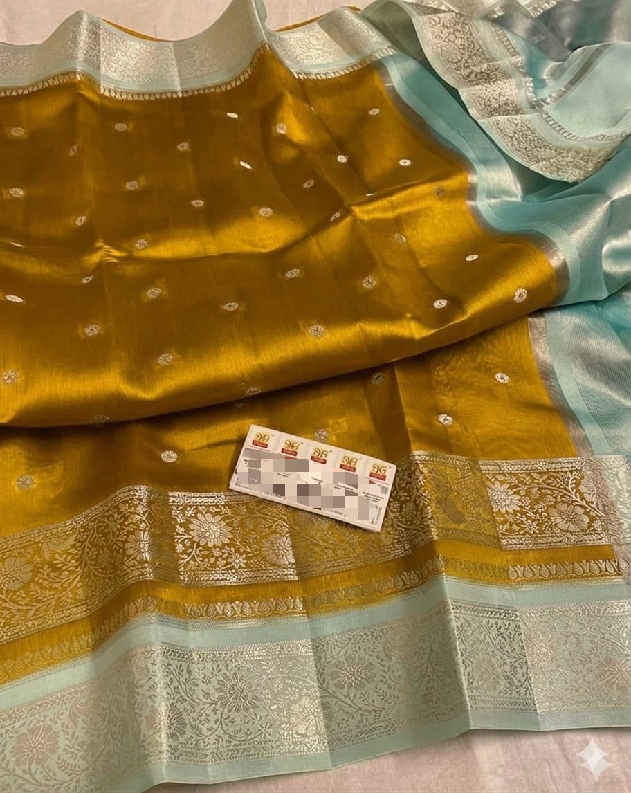A close-up of a draped fabric textile with a mustard yellow body, light aqua border, and ornate silver-toned woven borders. A small card is placed on the fabric.