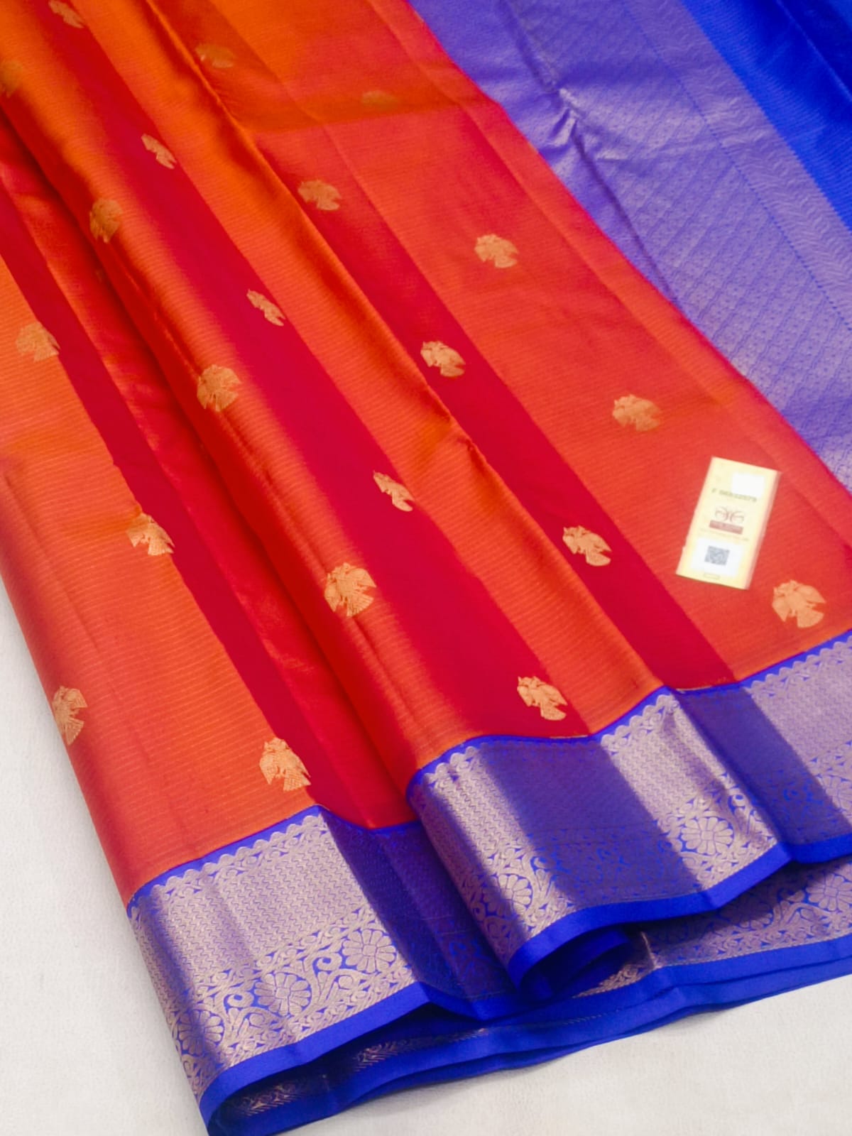 A close-up of a folded red and orange silk saree with gold zari motifs and a rich blue border.
