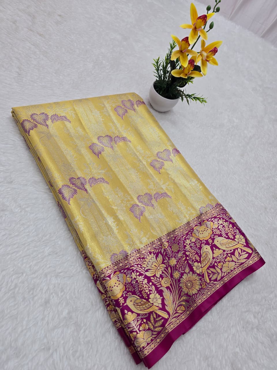 Yellow saree with purple border on a white surface, accompanied by a small potted plant.