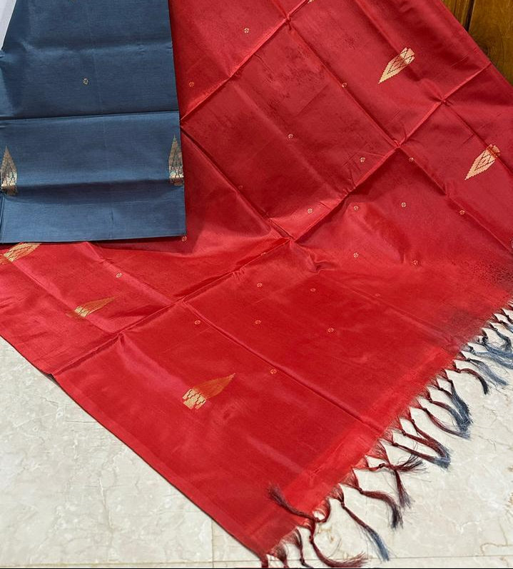 A red and navy blue silk saree with gold zari motifs and fringed edges, draped on a light-colored floor against a wooden door background.