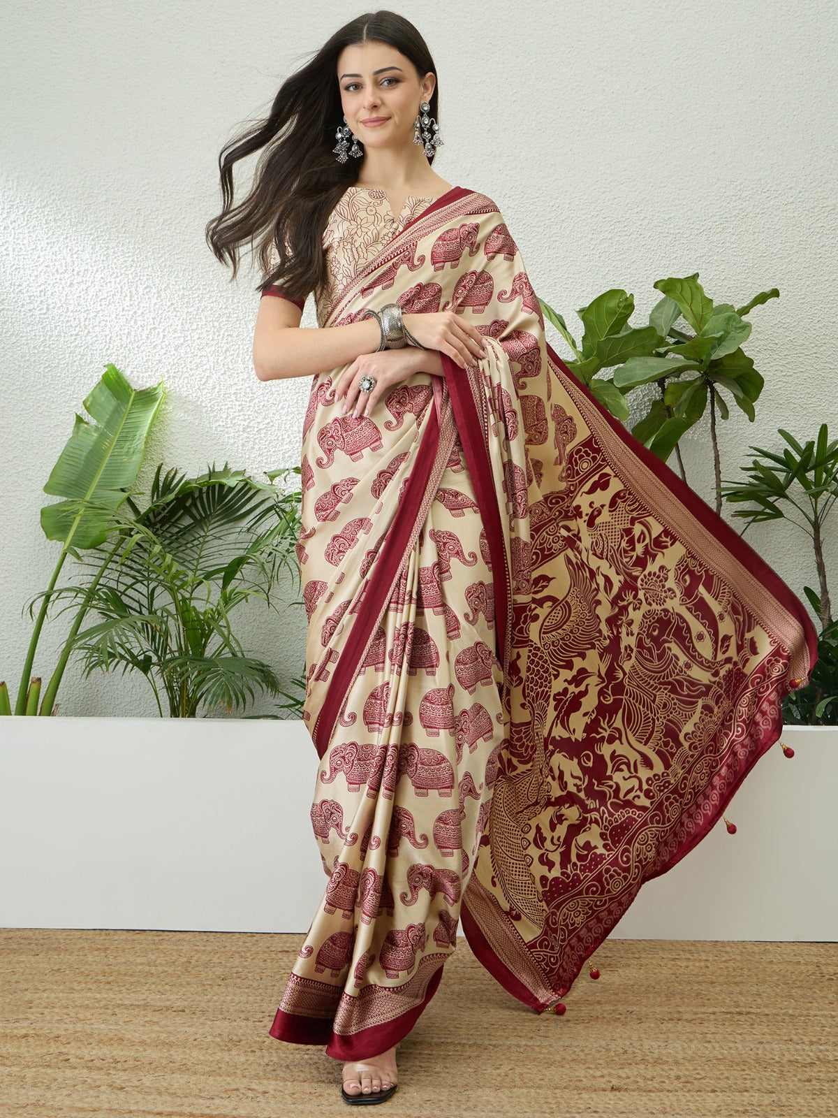 Woman wearing a traditional saree with a red and beige pattern, standing against a white wall with plants.