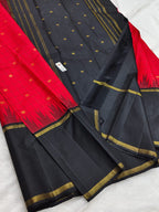 A black and red silk saree with gold zari borders and motifs, laid out on a white textured surface.
