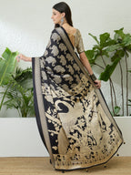 Woman wearing a black and gold saree with elephant patterns, standing against a white wall with green plants.