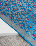 A close-up of a bright blue fabric with intricate silver floral embroidery and repeating pink and red paisley motifs, draped over a white surface with a gray shag rug underneath.