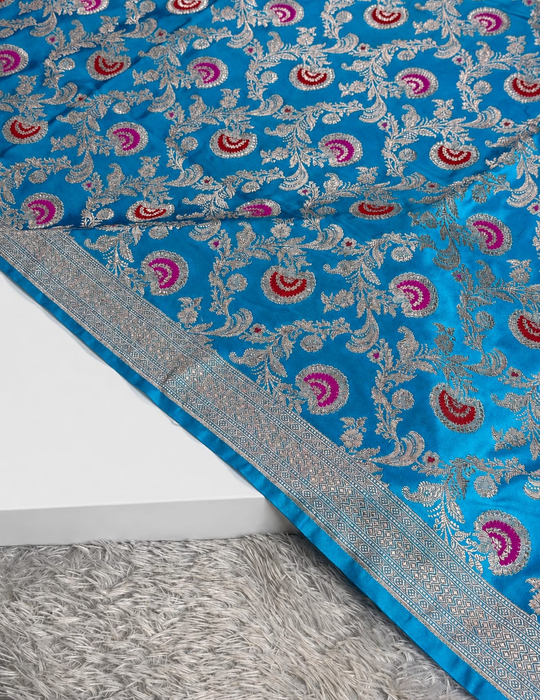A close-up of a bright blue fabric with intricate silver floral embroidery and repeating pink and red paisley motifs, draped over a white surface with a gray shag rug underneath.