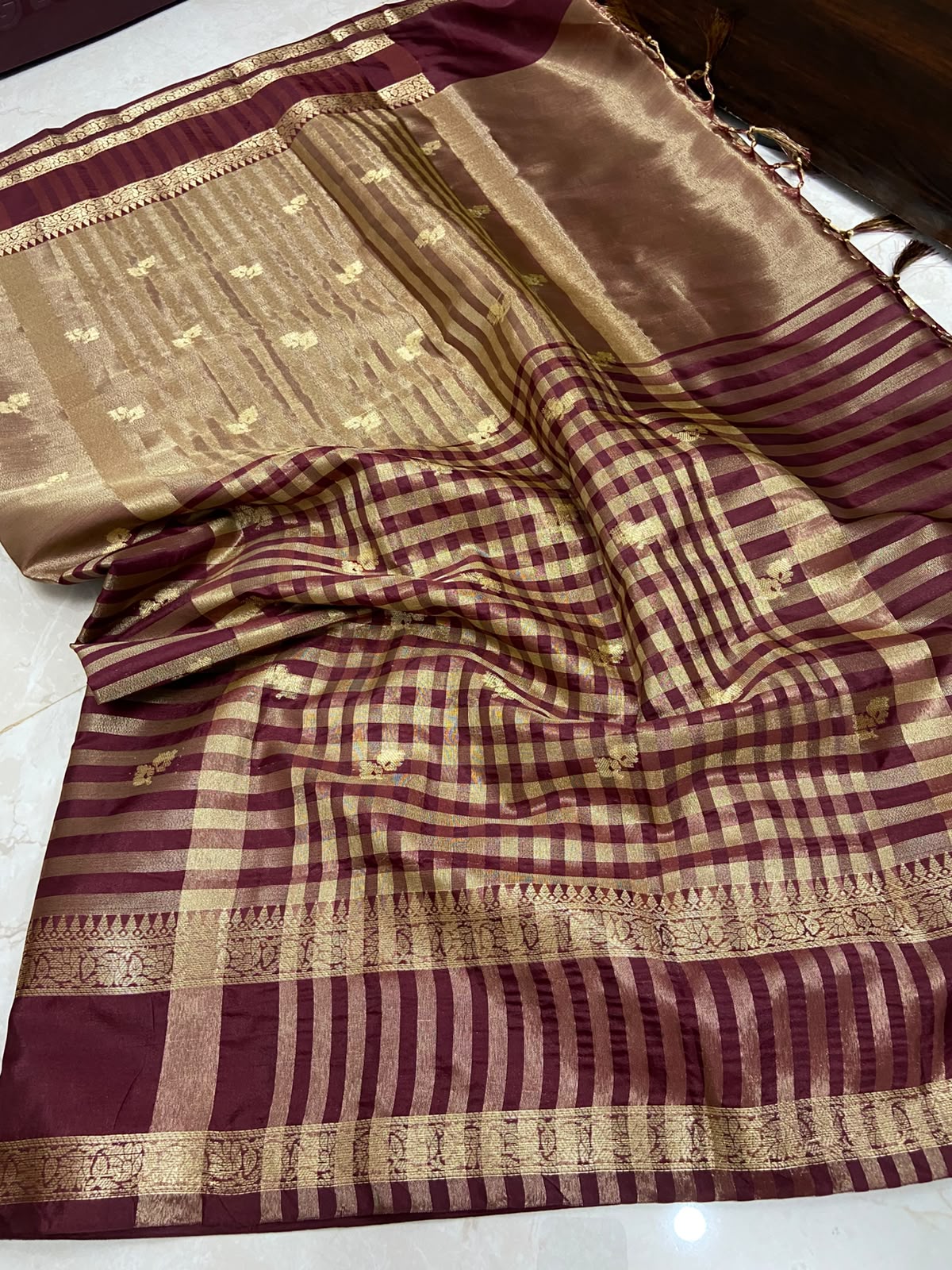 A draped silk saree with a deep maroon body, featuring a gold checkered pattern, a wide gold striped border, and a metallic gold pallu.