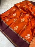 An orange silk saree with gold floral motifs and a deep maroon border, laid out on a light marble floor.
