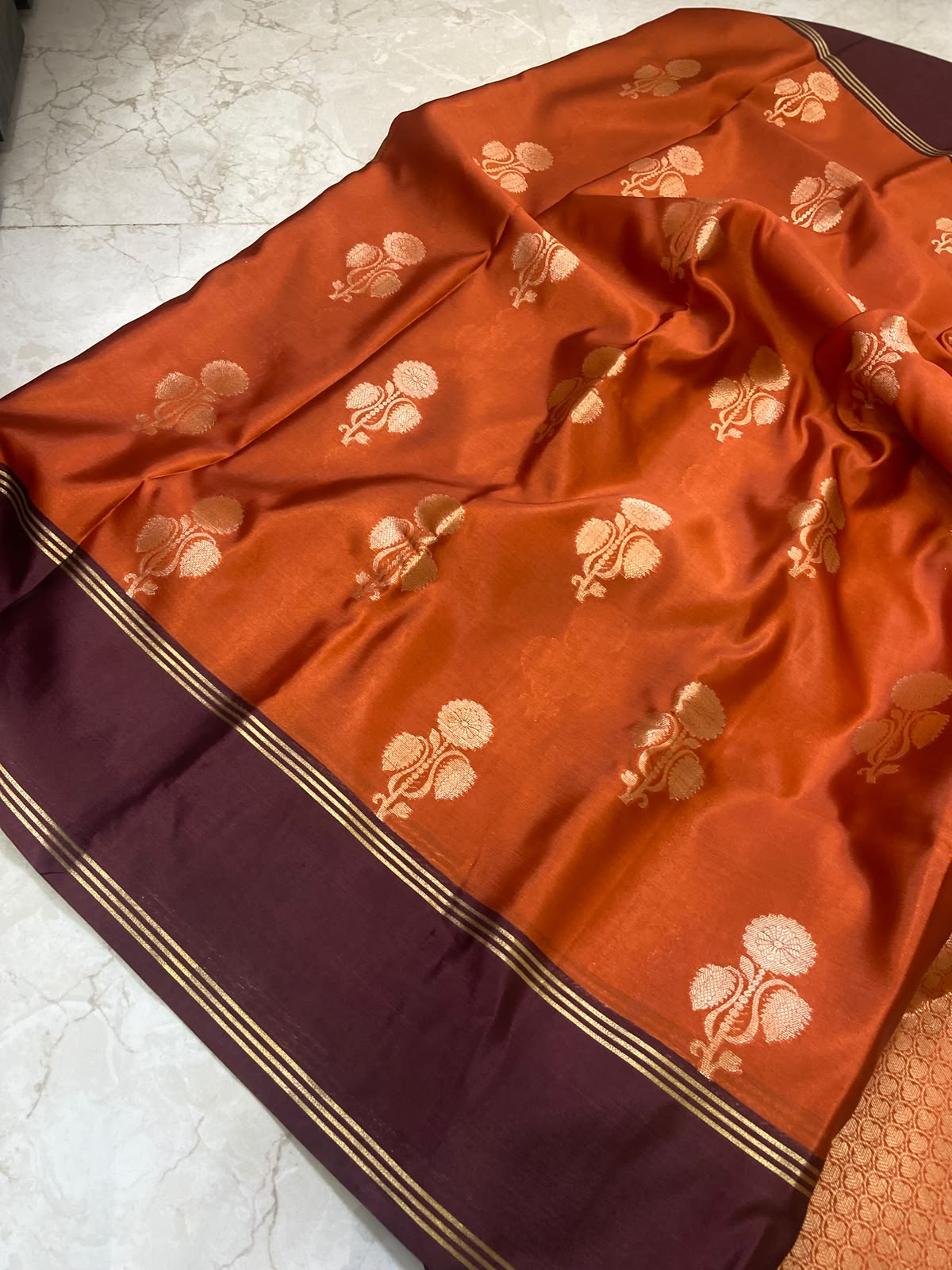 An orange silk saree with gold floral motifs and a deep maroon border, laid out on a light marble floor.