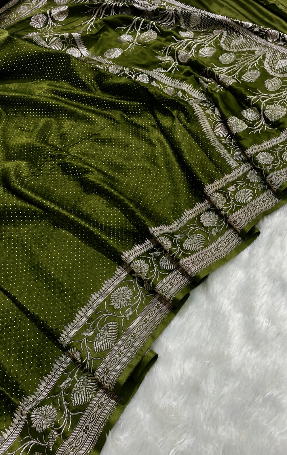 An olive green silk saree with intricate silver zari floral embroidery along its border, draped over a white textured surface.