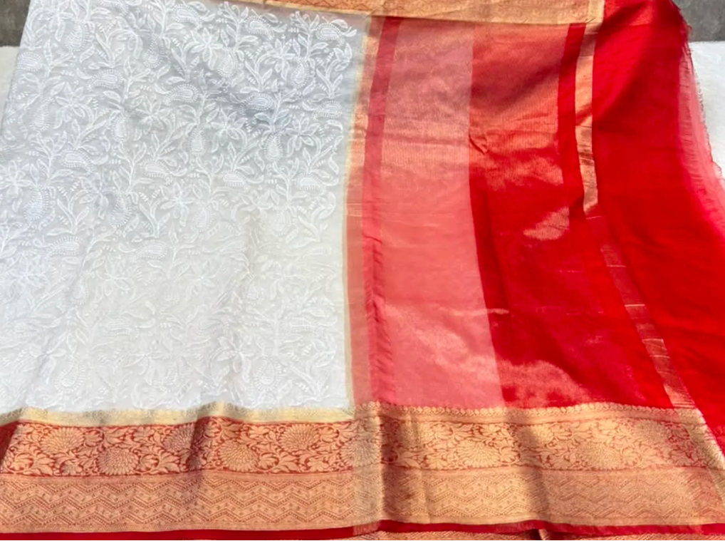 A white and red silk saree with intricate floral embroidery, a red pallu, and gold zari borders, held up by a person whose hands and torso are partially visible.