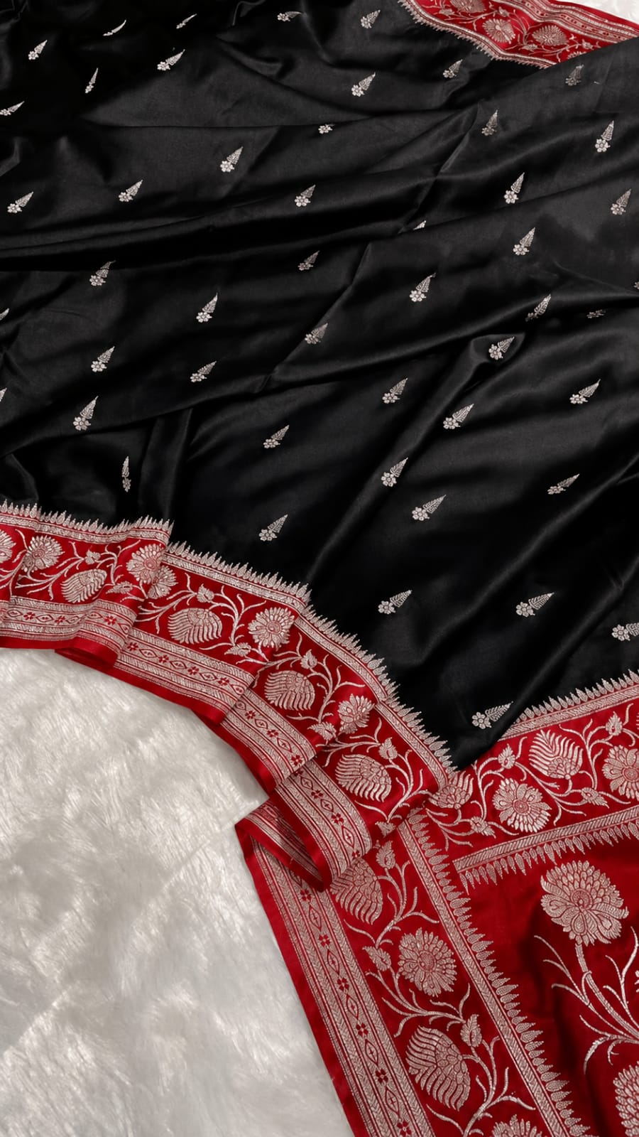 Black silk saree fabric with small silver motifs and a red border featuring intricate silver floral embroidery, laid over a white textured fabric.
