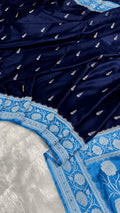 A close-up of a navy blue fabric with small silver motifs and a contrasting light blue border featuring intricate white floral embroidery.