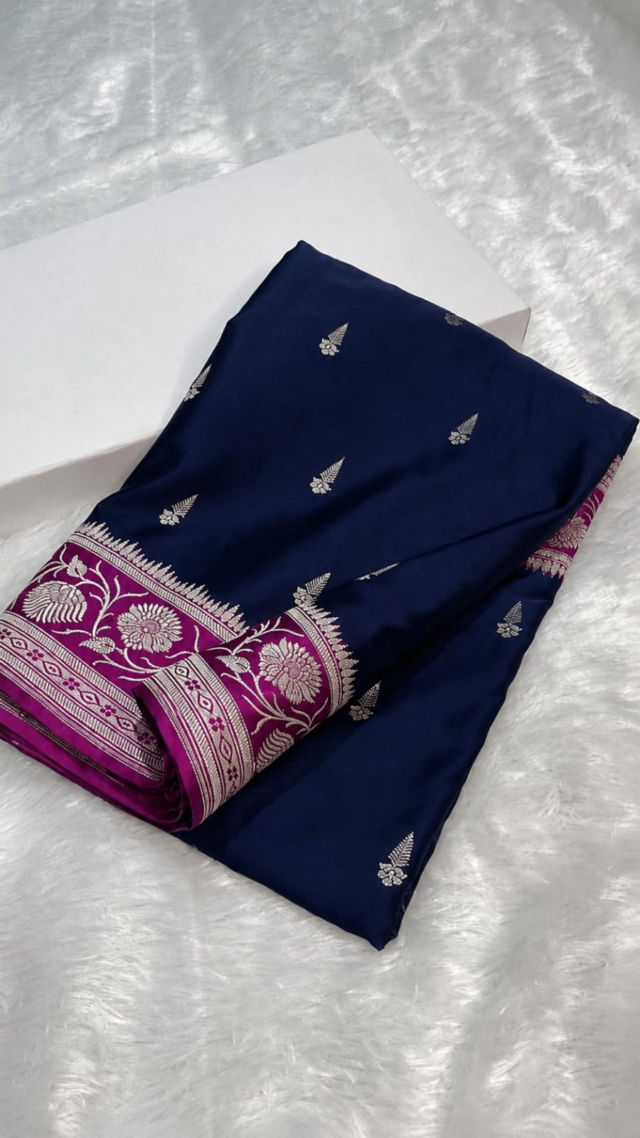 A folded navy blue fabric garment, likely a saree or dress material, featuring silver metallic motifs and a magenta border with intricate silver floral patterns, displayed on a white textured surface.