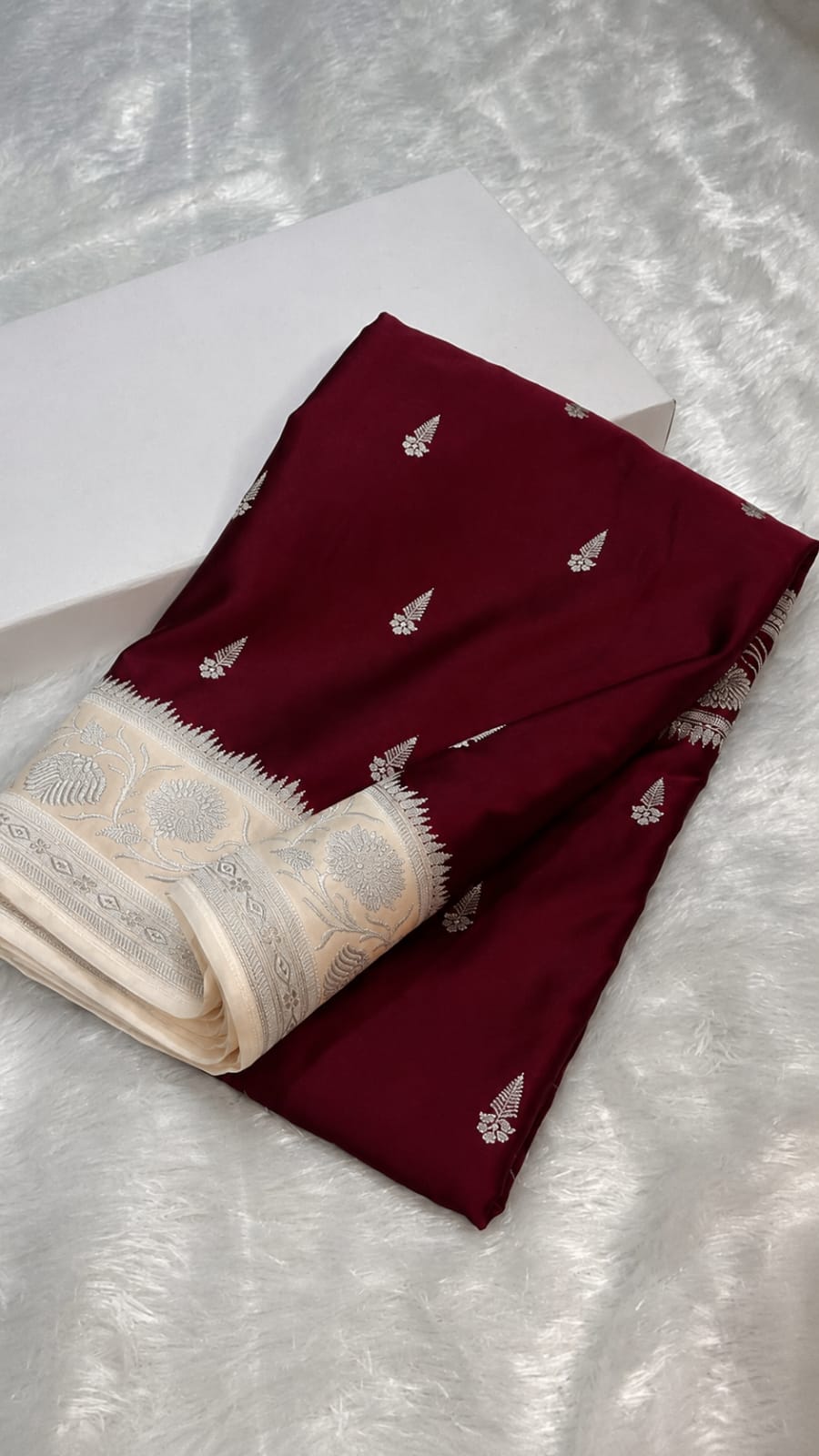 A folded deep maroon fabric piece with small silver motifs and a cream border, placed on a white textured surface next to a white box.