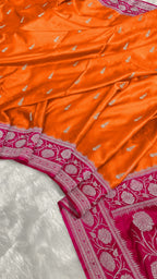 Bright orange Banarasi silk fabric with small silver zari motifs and a contrasting magenta border featuring intricate silver floral embroidery.