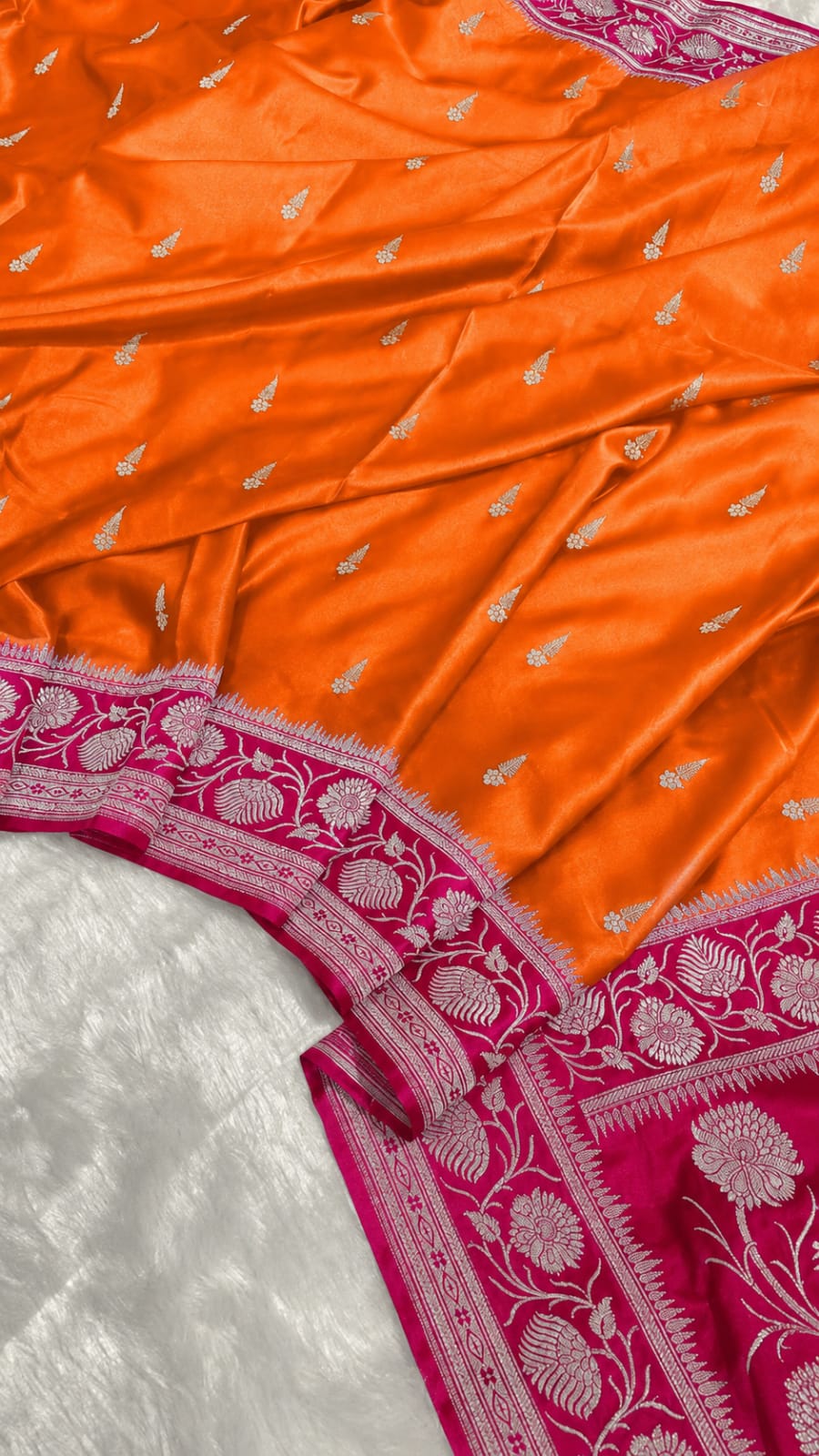 Bright orange Banarasi silk fabric with small silver zari motifs and a contrasting magenta border featuring intricate silver floral embroidery.