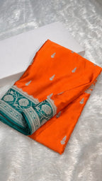 An orange fabric garment or textile piece with small silver motifs and a teal border featuring silver floral patterns, folded on a white textured surface next to a white box.