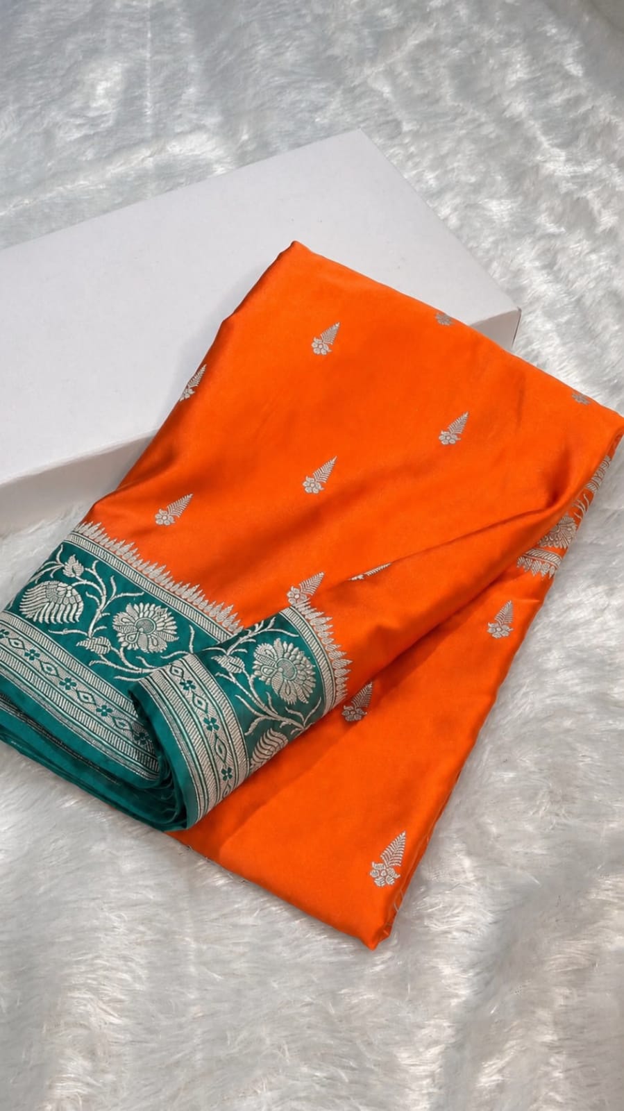 An orange fabric garment or textile piece with small silver motifs and a teal border featuring silver floral patterns, folded on a white textured surface next to a white box.