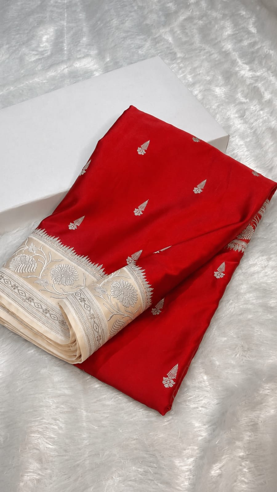 A folded red fabric garment or textile with small silver motifs and a wide cream border, placed on a white textured surface next to a white box.