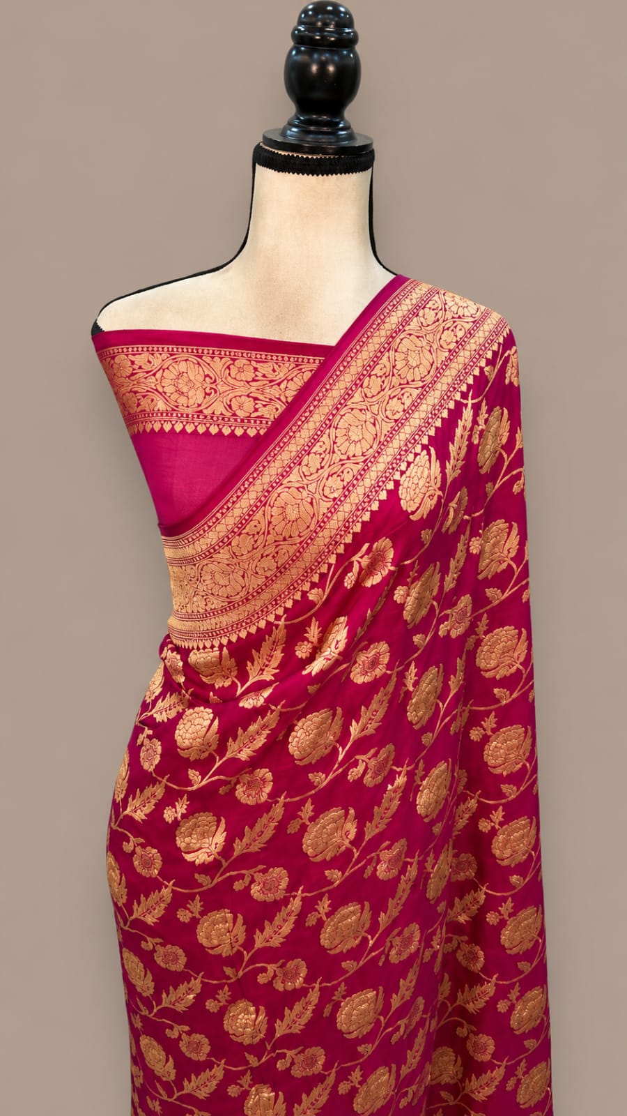A magenta pink silk saree with intricate gold zari floral brocade work, draped on a cream-colored mannequin torso against a solid taupe background.