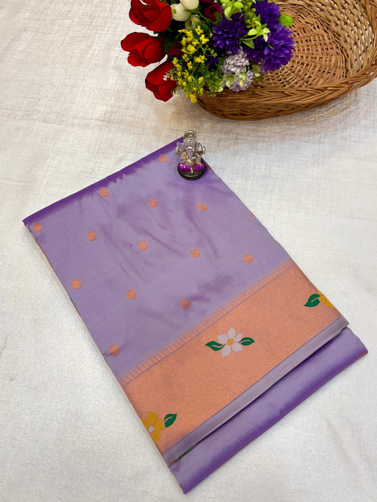 Purple and peach saree with floral patterns on a white surface, accompanied by a basket of flowers.