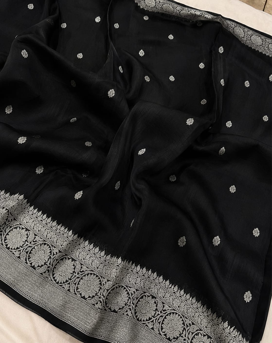 A black silk saree with silver zari embroidery, featuring small dot motifs scattered across the body and a wide decorative border with circular patterns.