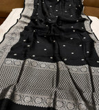 A black Banarasi silk saree with intricate silver zari embroidery, featuring a wide decorative border and small dot motifs, laid out on a white sheet.
