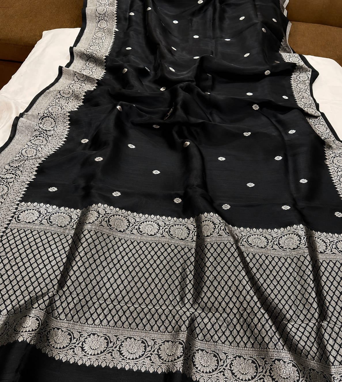 A black Banarasi silk saree with intricate silver zari embroidery, featuring a wide decorative border and small dot motifs, laid out on a white sheet.