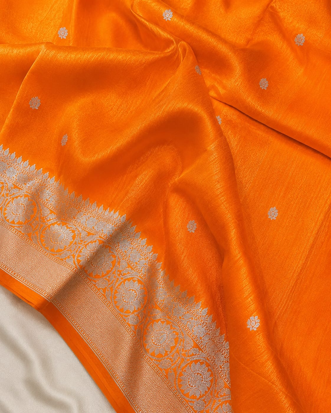 Bright orange silk saree with small silver floral motifs and a wide silver border with intricate zari work, draped elegantly over a white fabric.