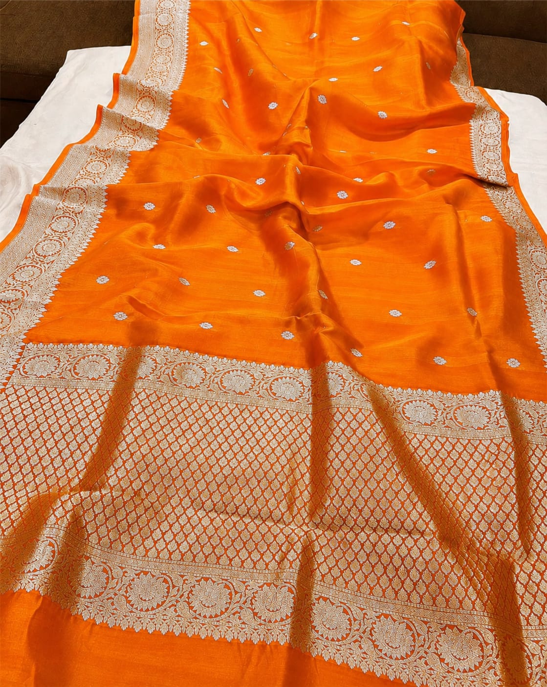 An orange silk saree with intricate white Banarasi zari embroidery, draped to display its border and body pattern.