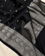 A close-up of a black fabric, likely Banarasi silk, featuring intricate silver zari woven patterns, including a detailed border and small scattered motifs.