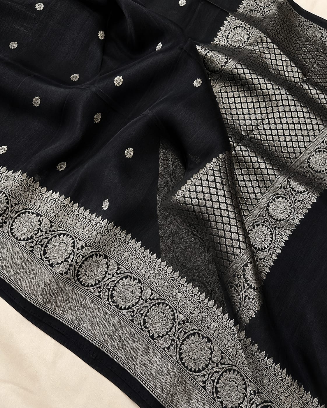 A close-up of a black fabric, likely Banarasi silk, featuring intricate silver zari woven patterns, including a detailed border and small scattered motifs.