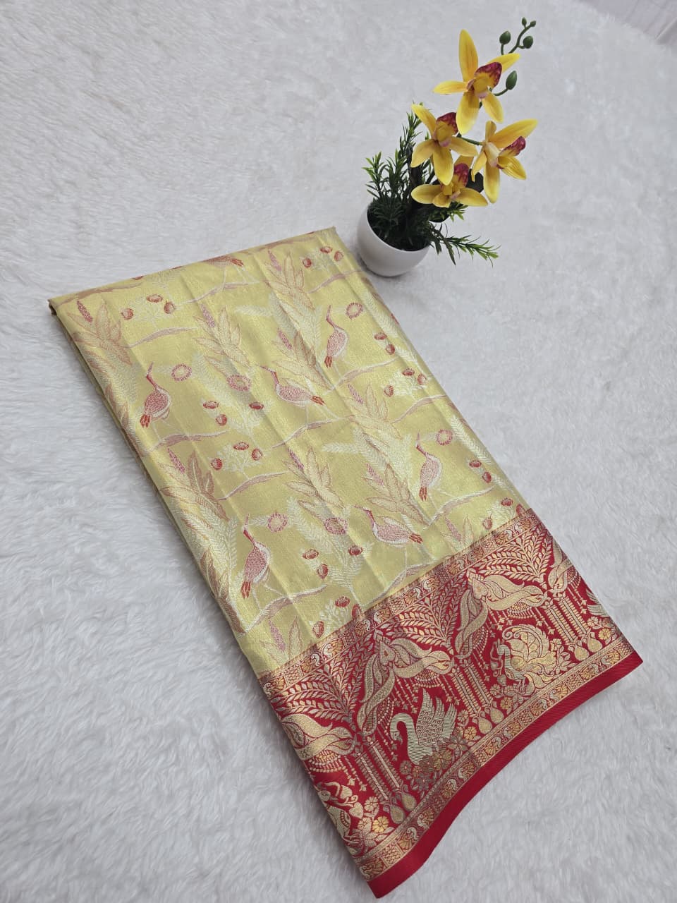 Decorative yellow and red fabric with floral patterns on a white surface, accompanied by a small potted plant with yellow flowers.