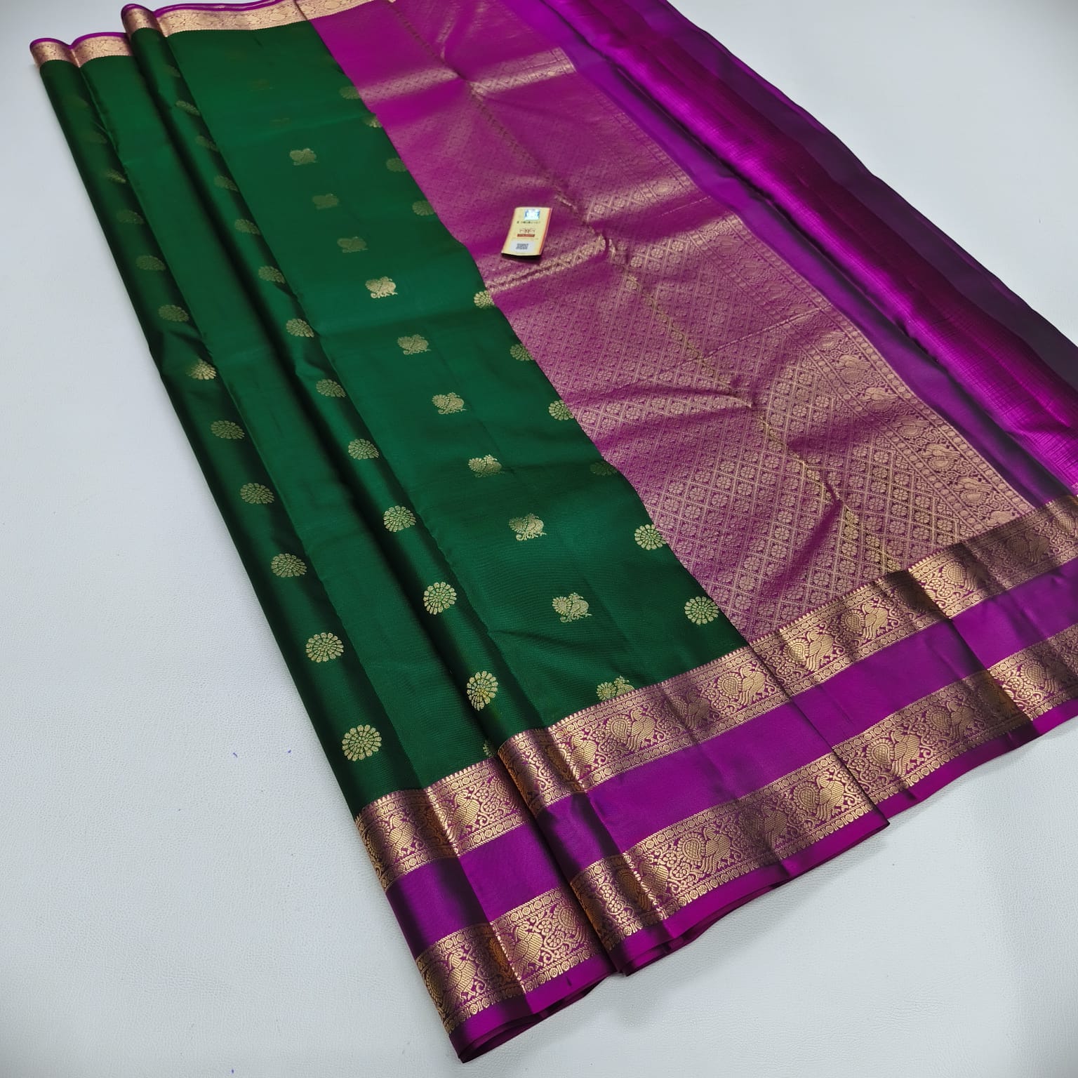 A dark green and magenta purple silk saree with intricate gold zari woven patterns and borders, laid out diagonally on a light gray background.