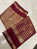 A folded silk saree displayed on a light beige marble surface, featuring a deep maroon border, a beige body with small gold motifs, and alternating maroon and gold stripes with a decorative gold zari border.