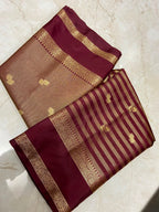 A folded silk saree displayed on a light beige marble surface, featuring a deep maroon border, a beige body with small gold motifs, and alternating maroon and gold stripes with a decorative gold zari border.